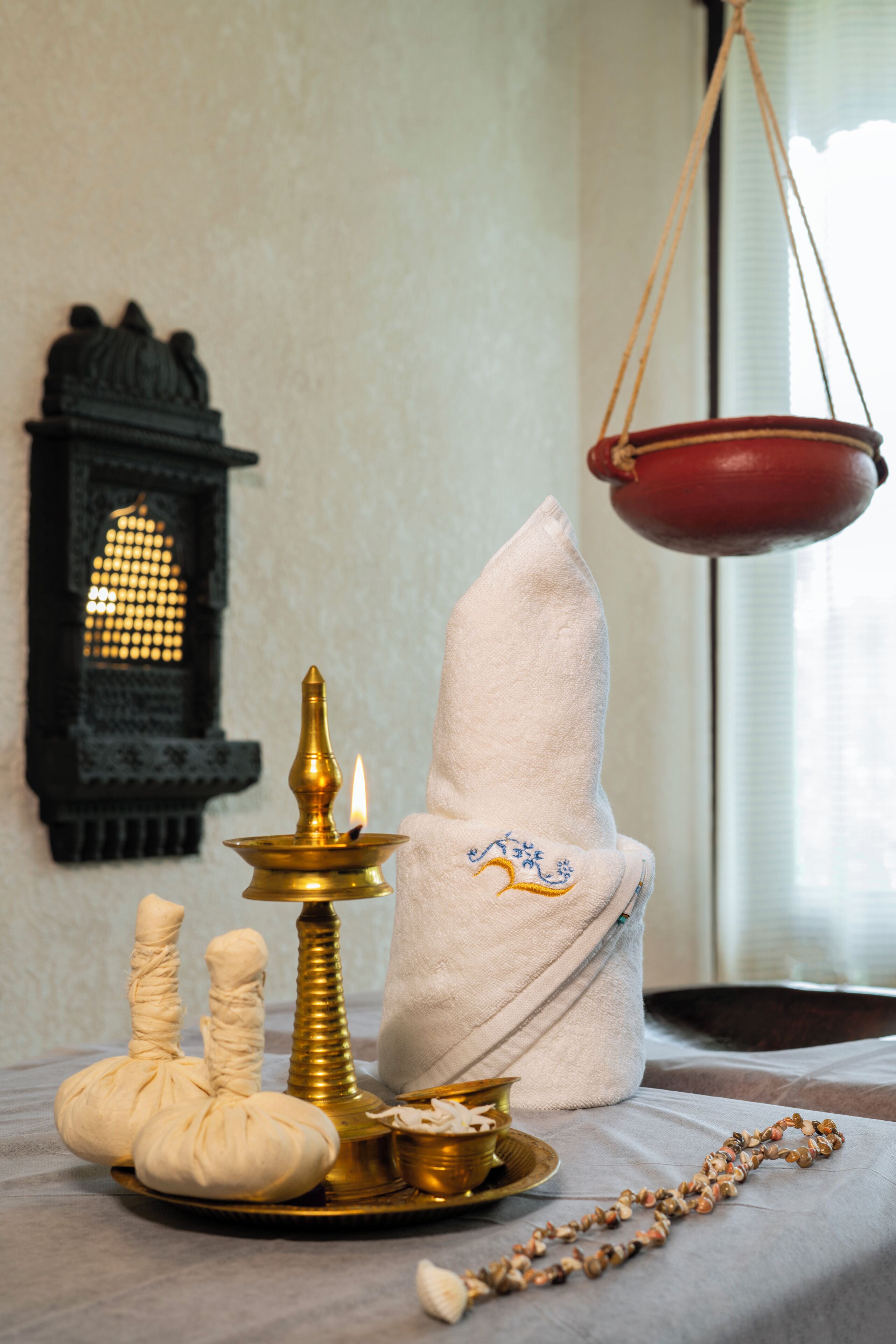 Une mise en scène traditionnelle avec une lampe à huile allumée, un pot rouge suspendu, un tissu blanc et un chapelet sur une table.