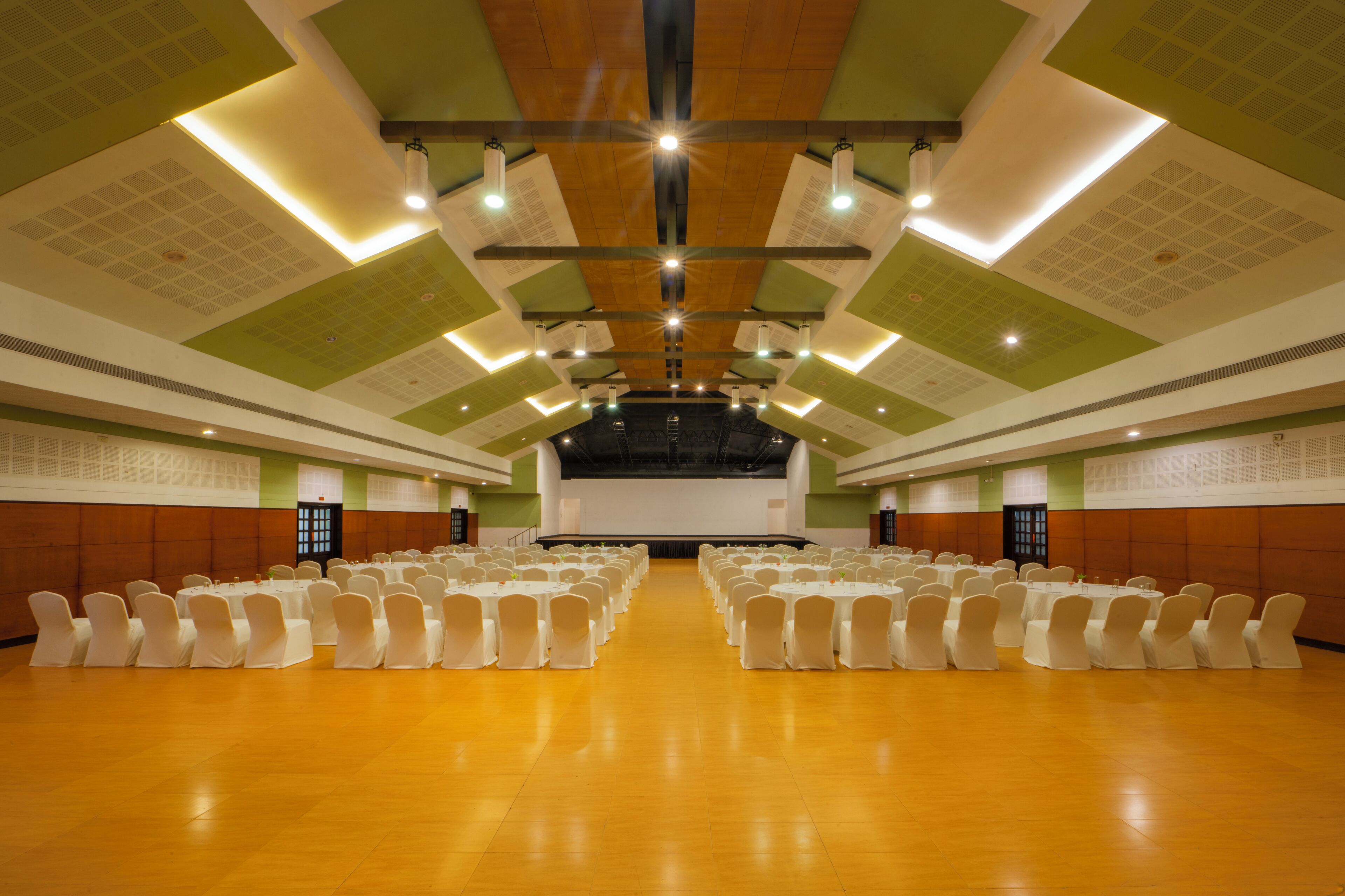 Een ruime zaal met rijen witte stoelen en tafels, een podium aan de voorzijde en een plafond met groene en witte patronen.