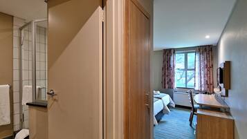 A hotel room with a wooden door, a bathroom with a shower, a bed with a white comforter, a desk with a mirror, and a window with curtains.