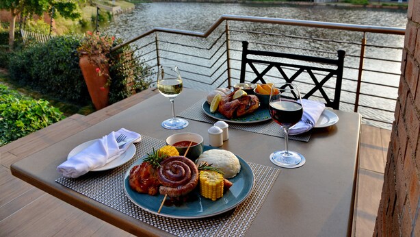 A dining table with a meal, wine glasses, and a view of a river and buildings. - Town Lodge Roodepoort (Roodepoort)