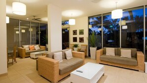A modern living room with wicker furniture, a white coffee table, and large windows with a view of palm trees. - Whitsunday Apartments (Whitsundays)