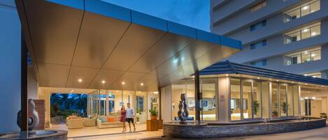 A modern hotel entrance with a covered outdoor seating area, a reception desk, and a large glass window.