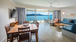 A modern living room with a dining area, a sofa, and a TV. It features large windows with a view of the sea and mountains. - Whitsunday Apartments (Whitsundays)