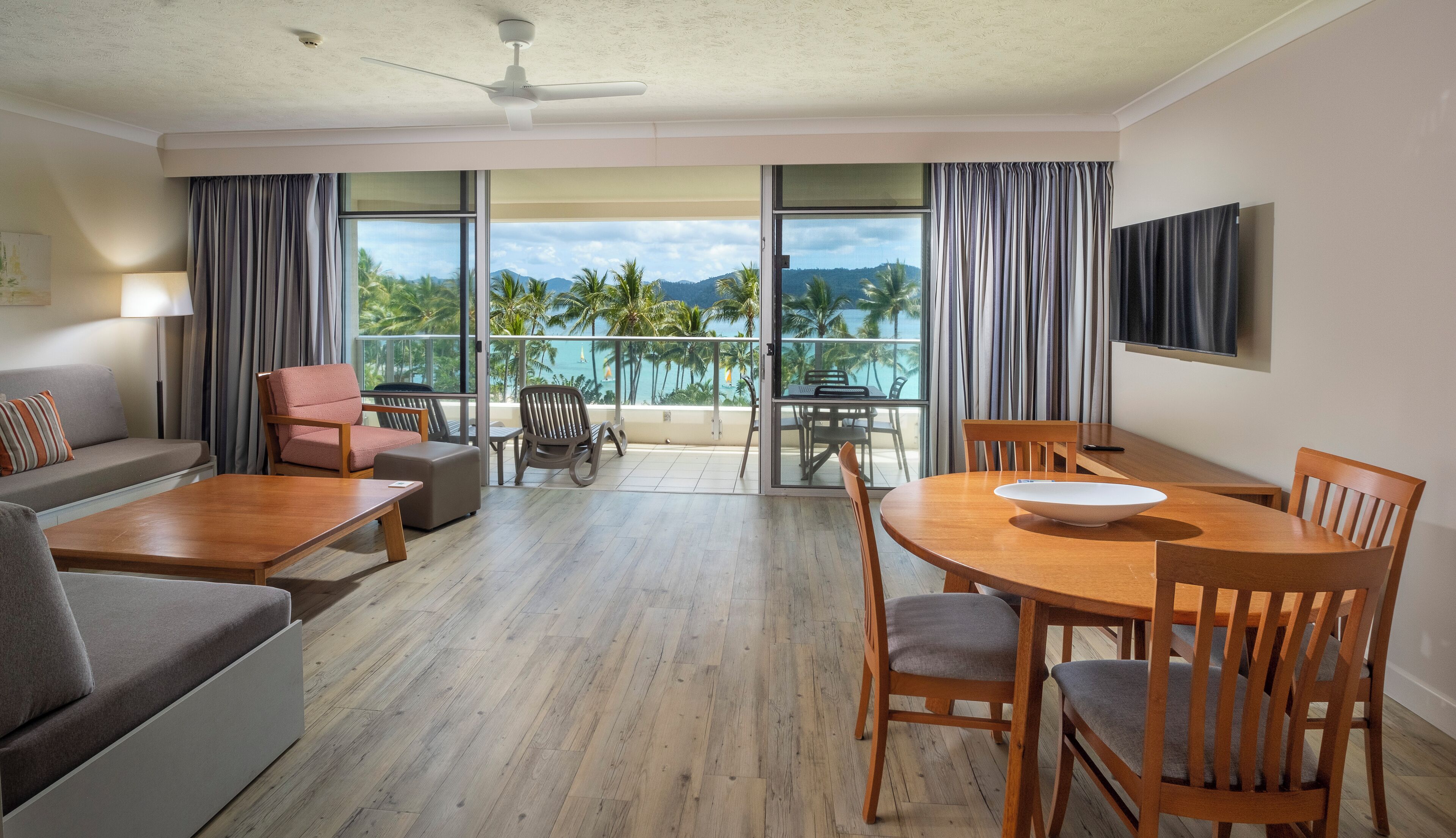 A modern hotel room with a balcony, a sofa, a dining table, and a TV mounted on the wall.