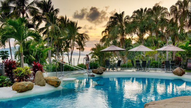 A pool area with lounge chairs and umbrellas, surrounded by palm trees and rocks. - Whitsunday Apartments (Whitsundays)
