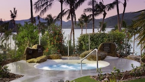 A spa area with a hot tub surrounded by tropical plants and palm trees, overlooking a body of water. - Whitsunday Apartments (Whitsundays)