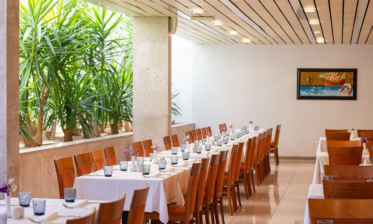 A long dining table with chairs, white tablecloths, and glassware, set in a room with a tiled floor and a painting on the wall.