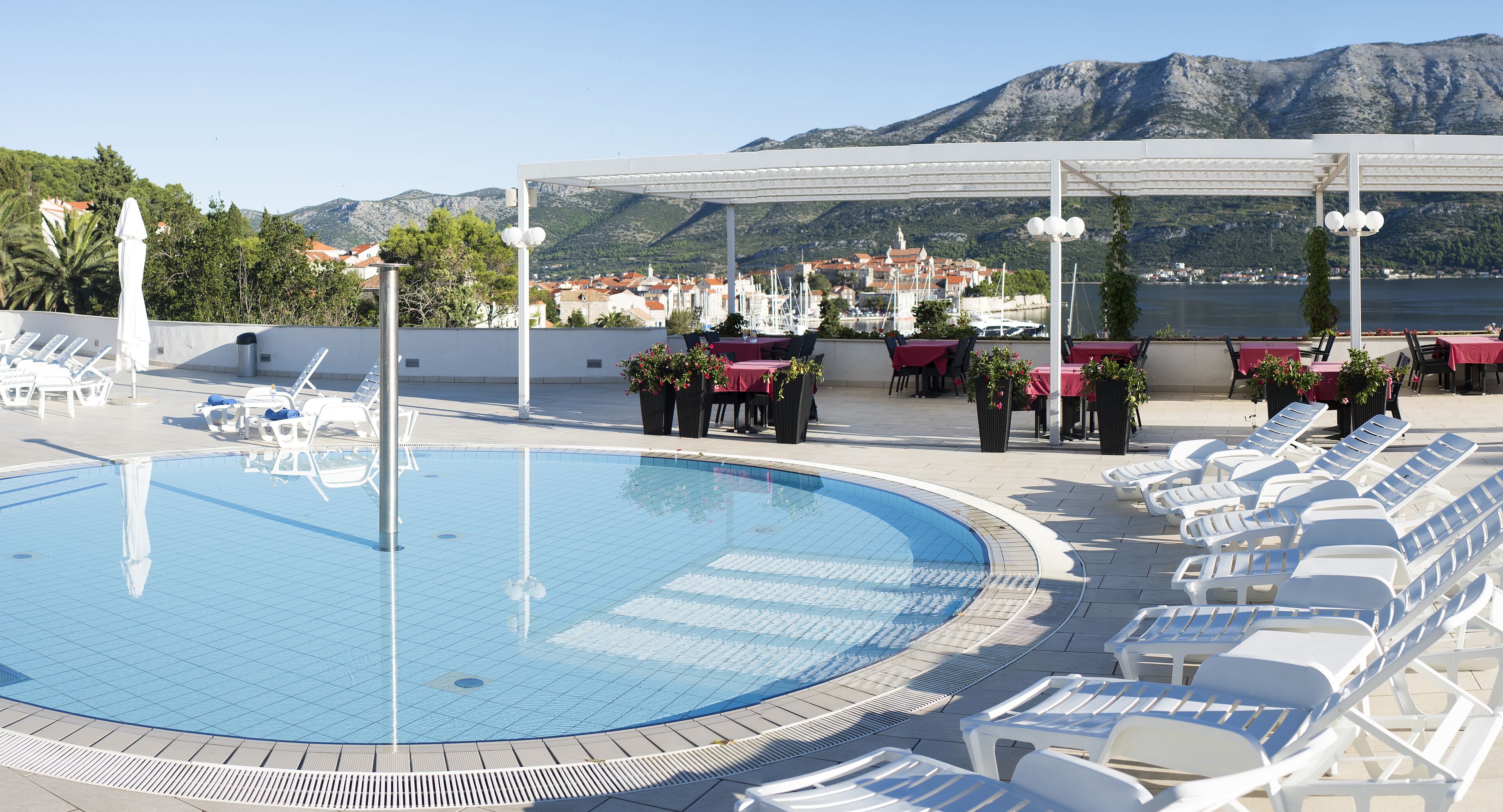 Area piscina con lettini, tavoli, una pergola e vista su una città e montagne sullo sfondo.