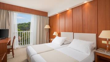 Ein Hotelzimmer mit zwei Betten, einem Schreibtisch, einem Fernseher und einem Balkon mit Vorhängen.