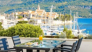A terrace with a table, chairs, and a glass of orange juice. In the background, there is a marina with boats and a coastal town with buildings and a hill.