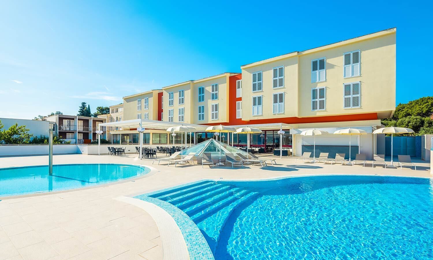 A swimming pool area with lounge chairs and umbrellas, surrounded by multi-story buildings with balconies.