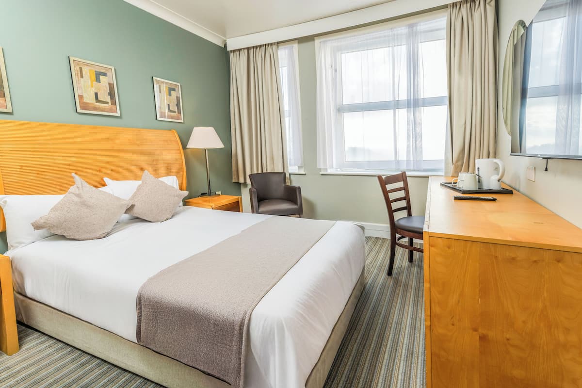 A hotel room with a large bed, a desk, a chair, a lamp, and a window with curtains.
