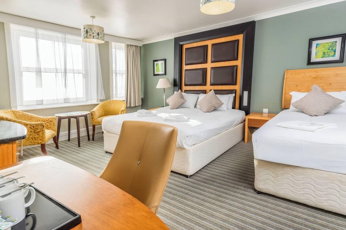 A hotel room with two beds, a dining table, a chair, a lamp, and a window with curtains.