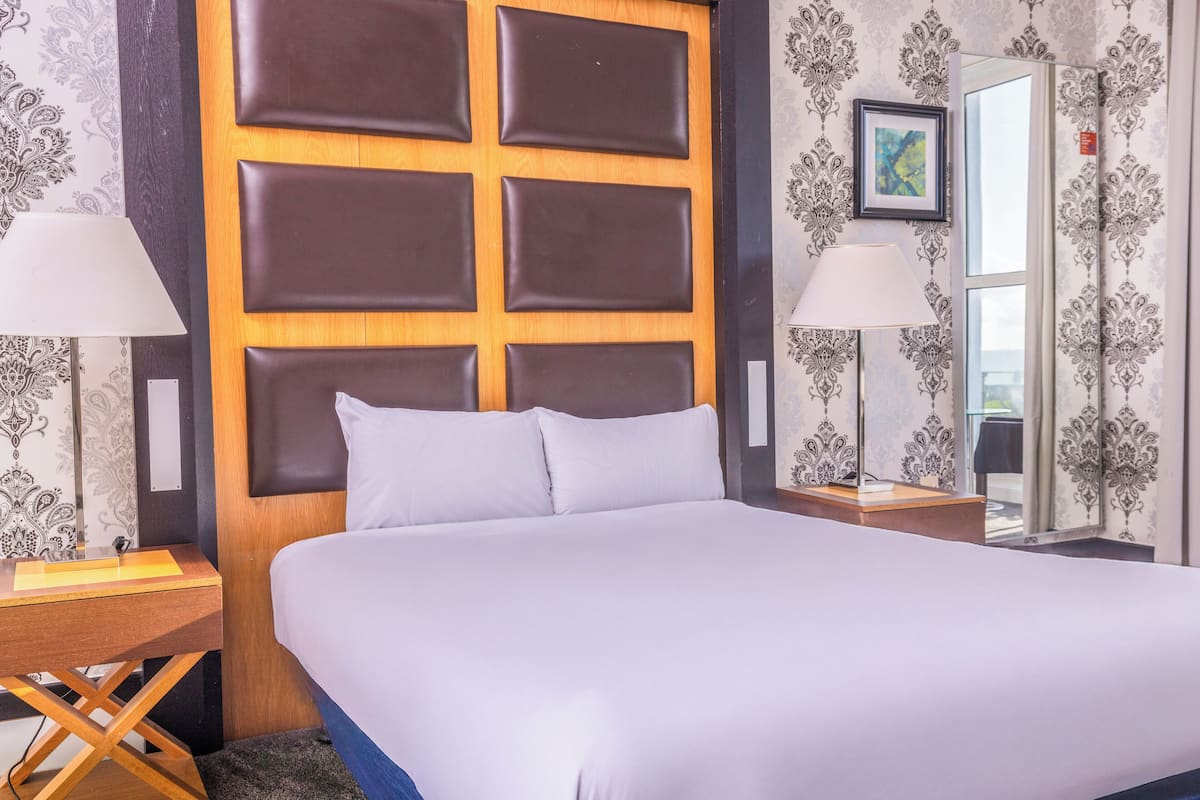 A hotel room with a large bed, wooden headboard, two bedside tables with lamps, and patterned wallpaper.