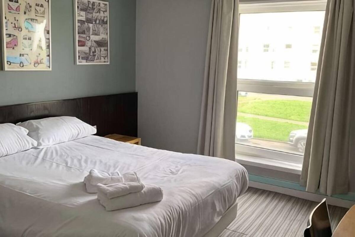 A hotel room with a bed, a window with curtains, and artwork on the wall.