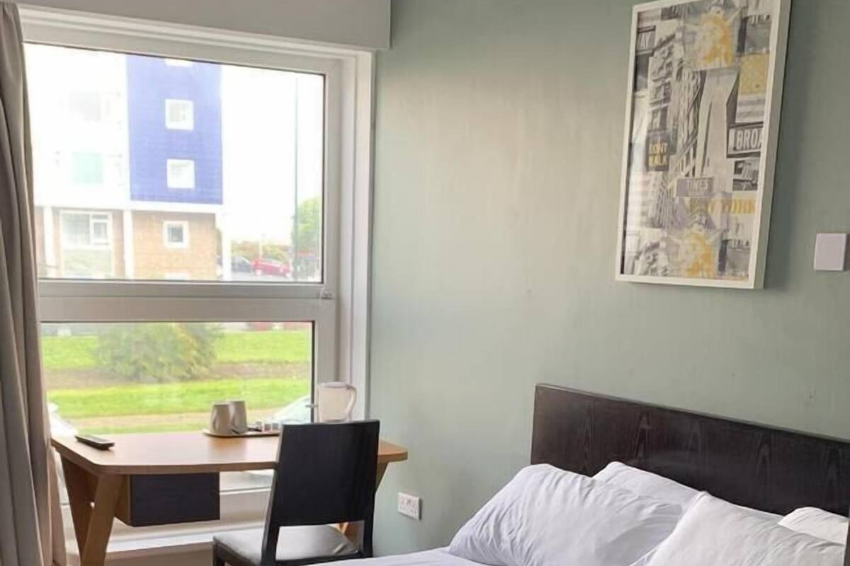 A hotel room with a bed, a desk, and a chair. There is a window with a view of buildings outside.