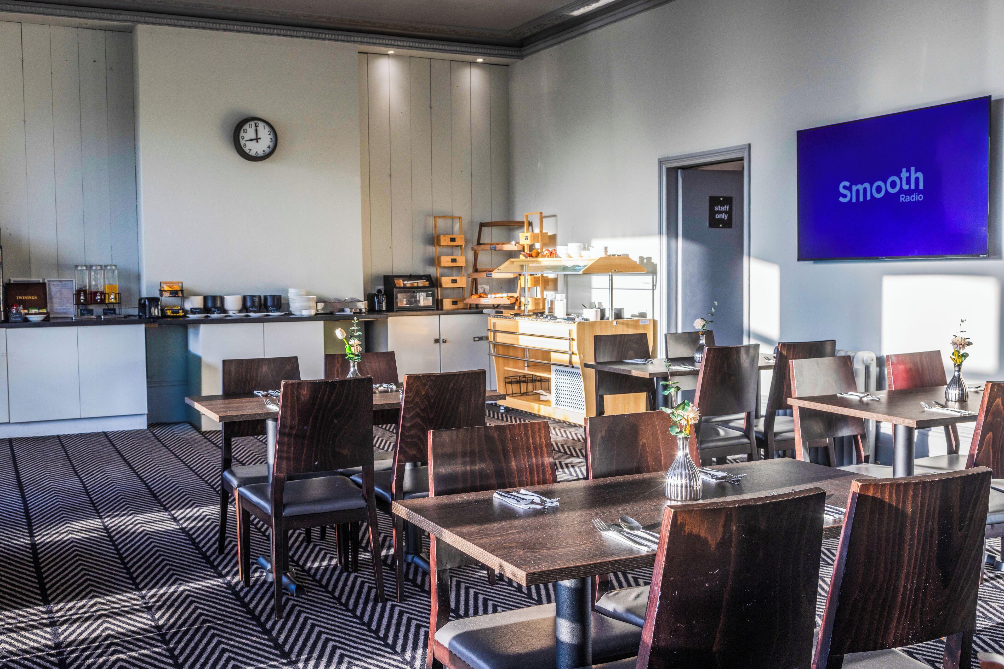 A modern restaurant with wooden tables and chairs, a kitchen area, and a clock on the wall.