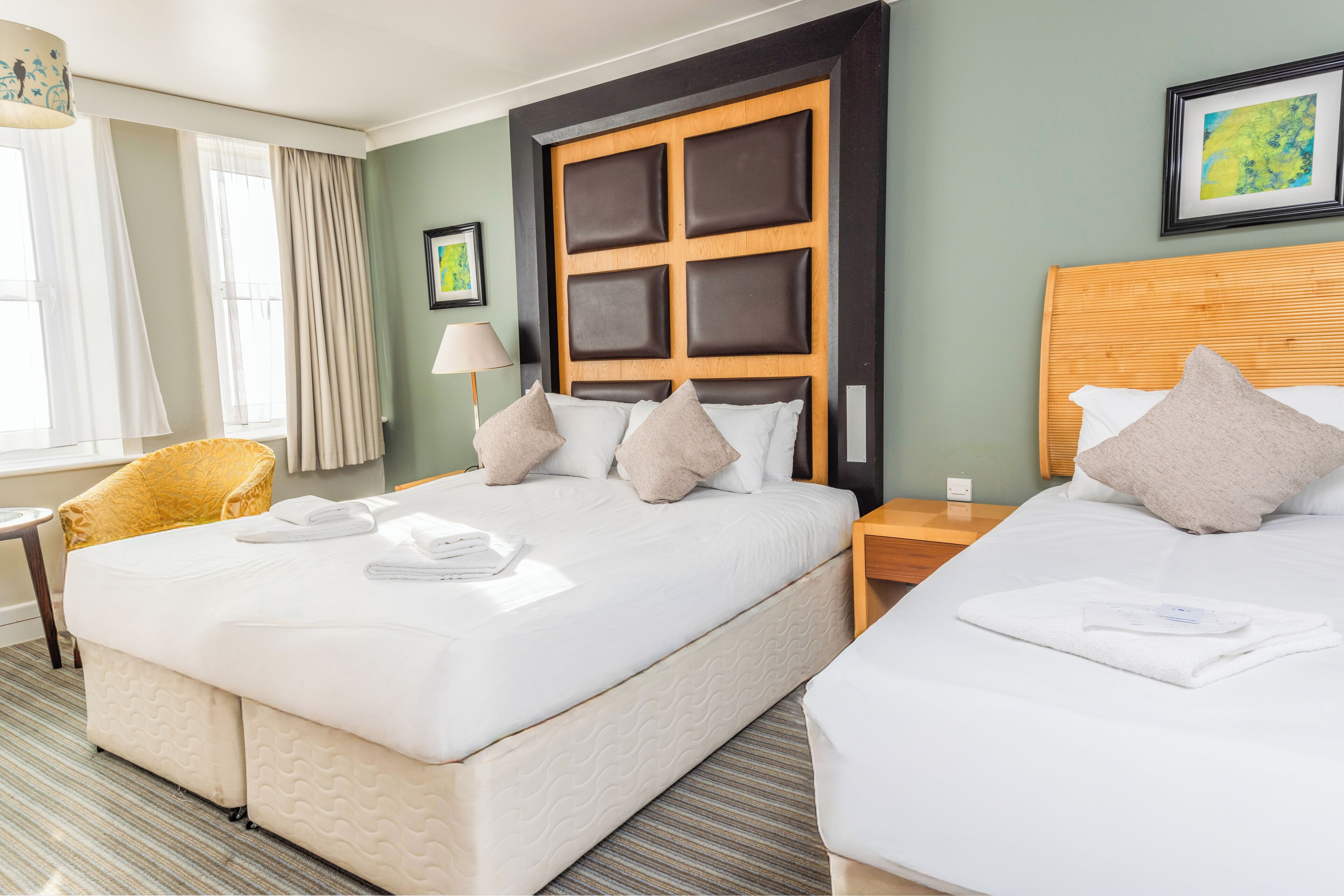 A hotel room with two beds, a wooden headboard, a yellow armchair, and a window with curtains.