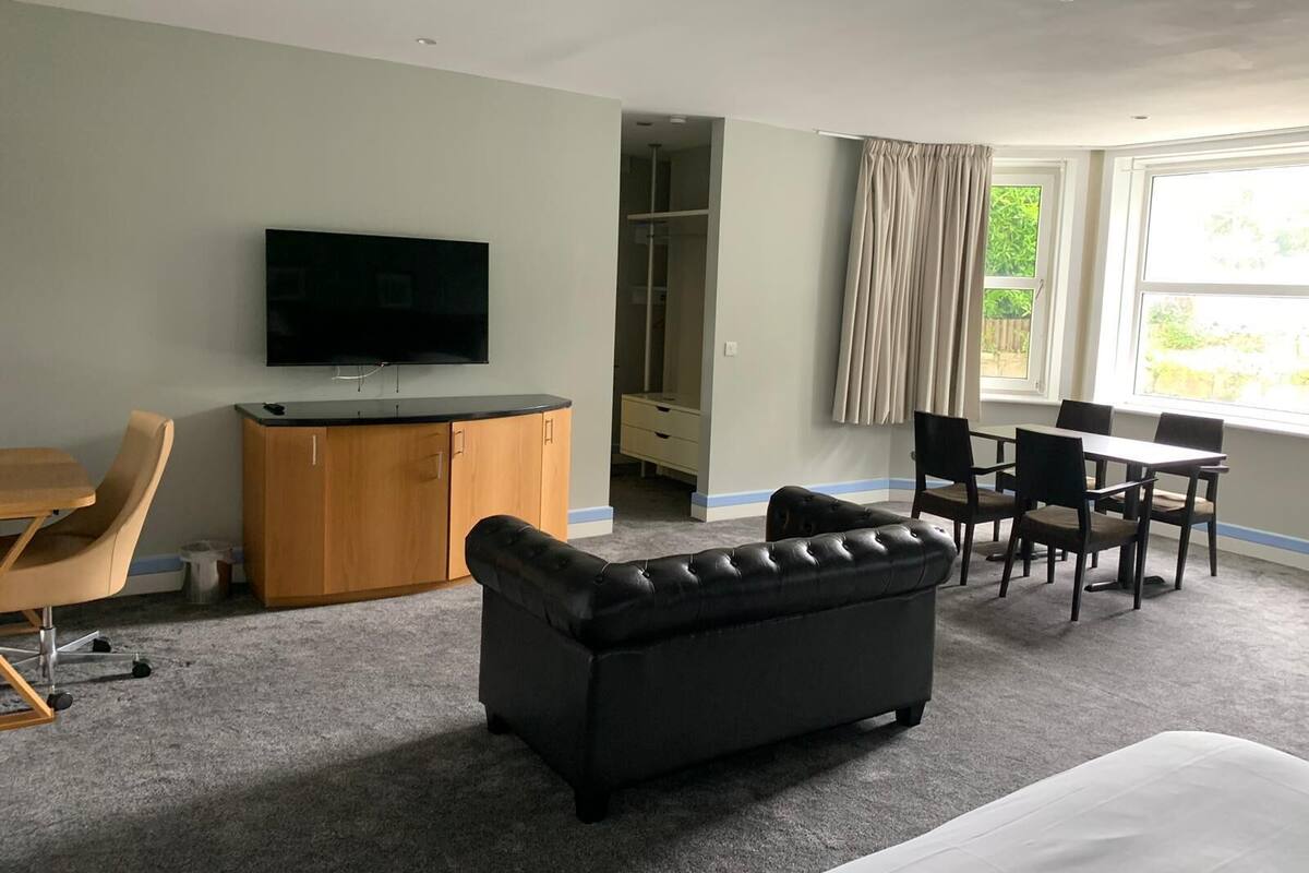 A hotel room with a flat-screen TV, a sofa, a dining table with chairs, and a window with curtains.