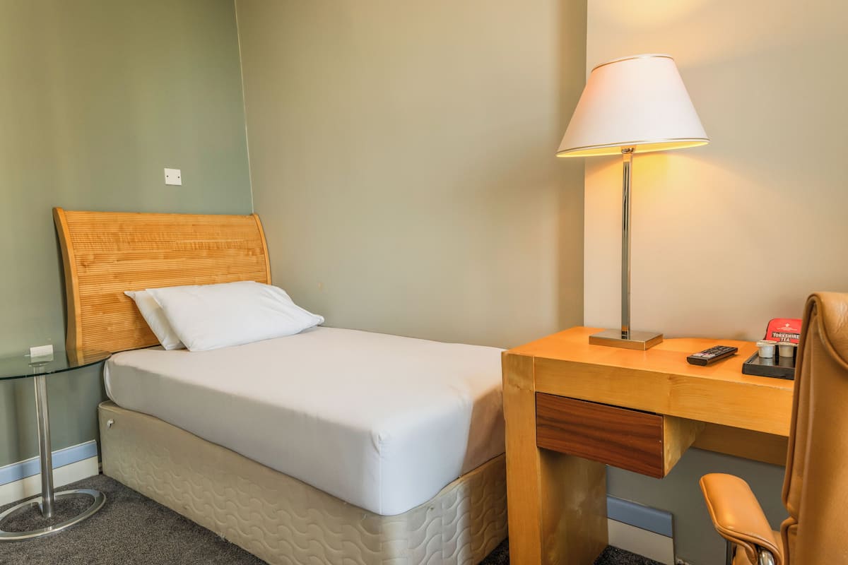 A neatly arranged hotel room with a single bed, a desk with a lamp, a chair, and a small table.