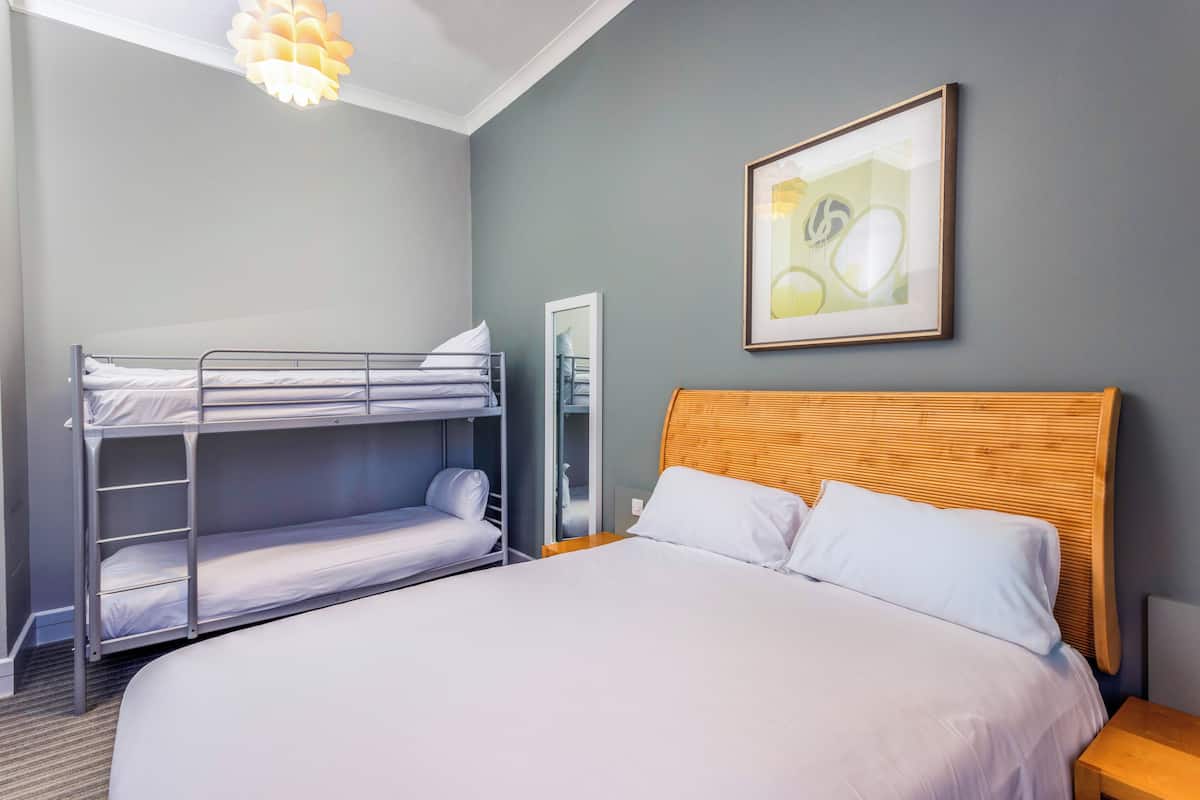 A hotel room with a bunk bed, a single bed, a wooden headboard, a framed picture on the wall, and a bedside table.
