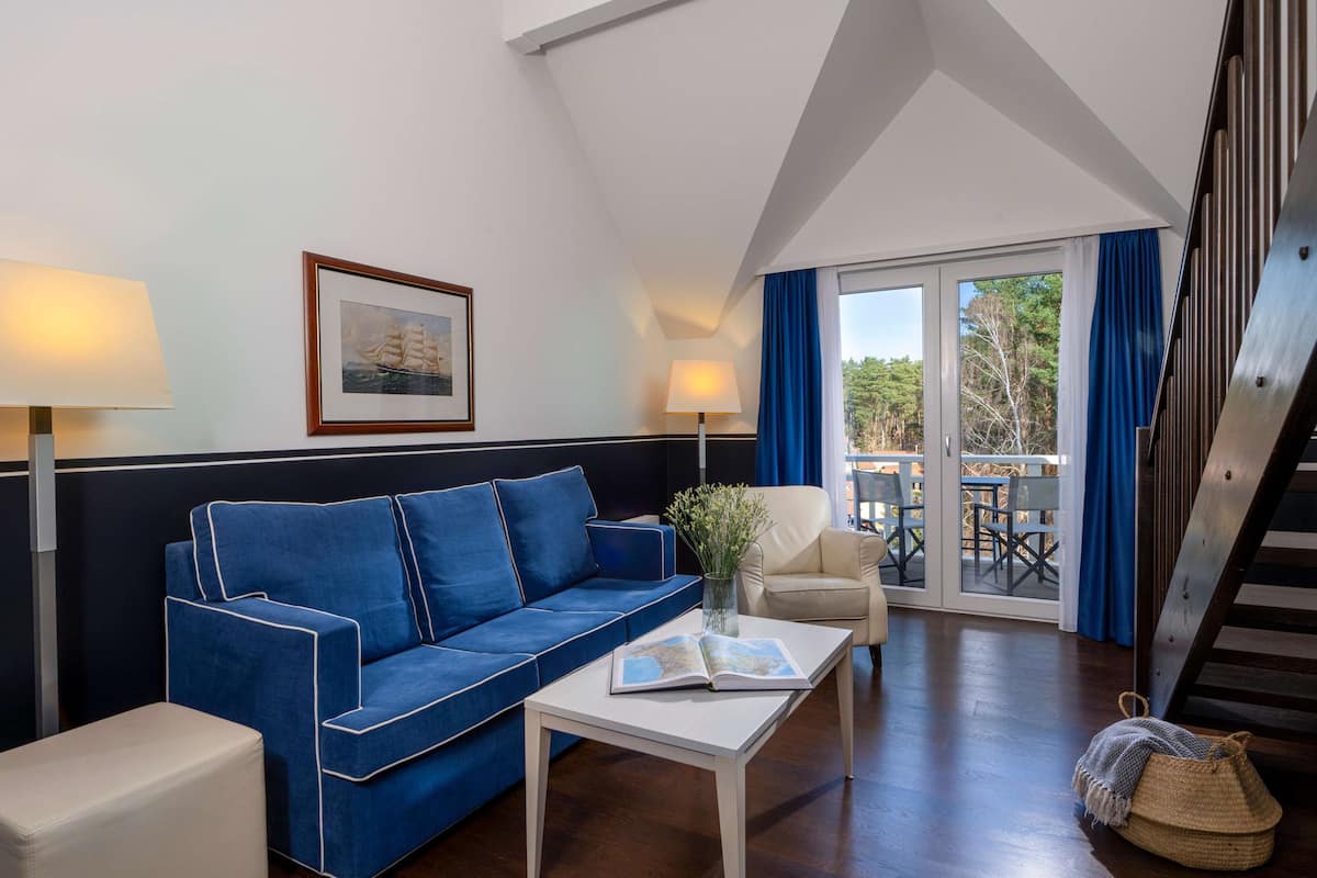 Un salon moderne avec un canapé bleu, une table basse blanche et un fauteuil beige. Un tableau encadré est accroché au mur, il y a une lampe, et on peut voir l’extérieur à travers une porte vitrée.