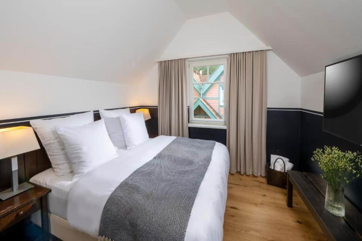 A hotel room with a bed, a TV, a window with curtains, and a view of buildings outside.