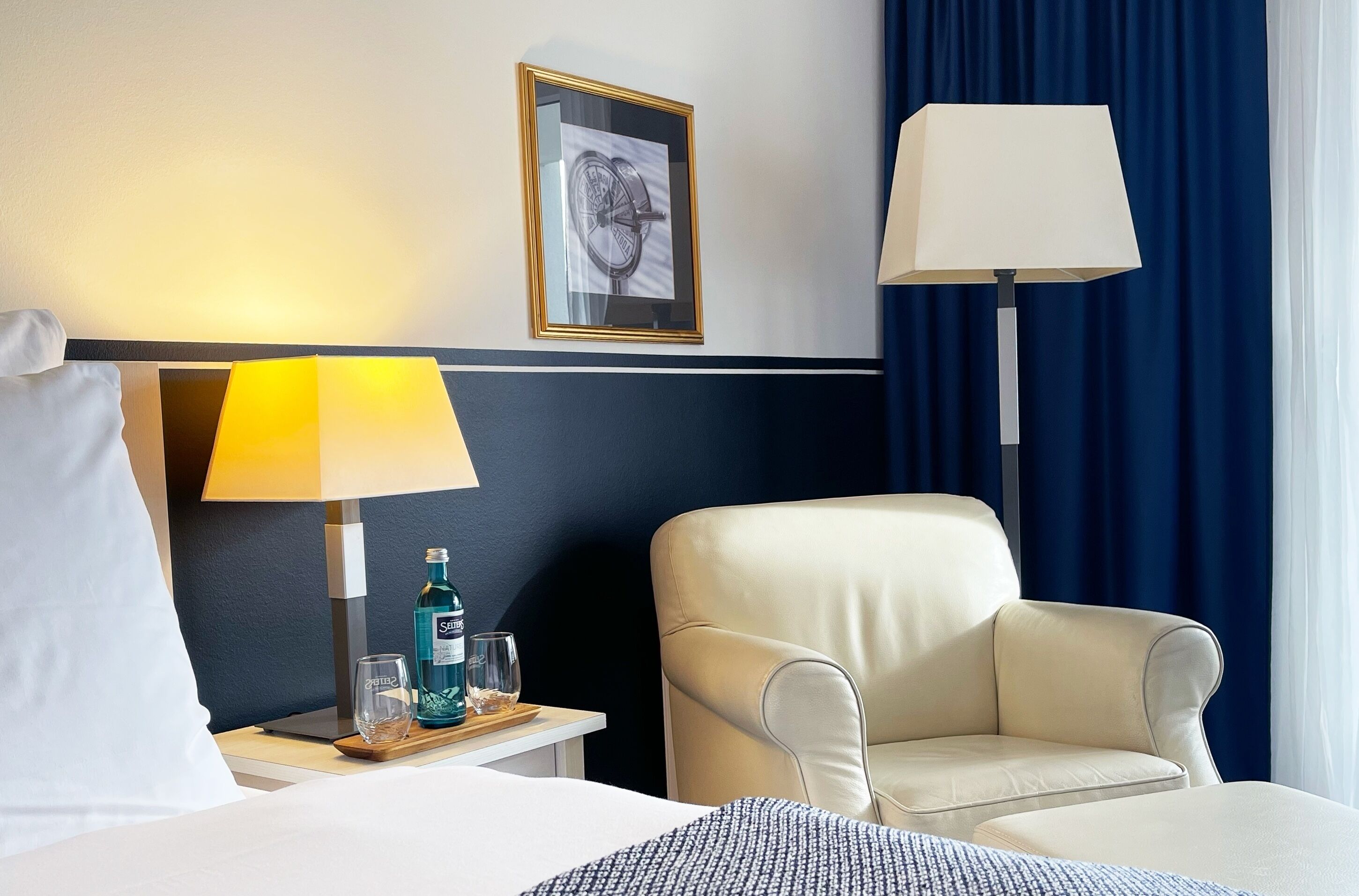 A hotel room with a bed, a nightstand with a lamp, a chair, a framed picture on the wall, and a window with curtains.
