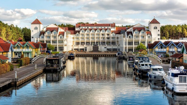 A waterfront hotel with multiple balconies and a central tower, boats docked in the water, and a pedestrian bridge connecting the buildings. - Sunday Resort Hafendorf Rheinsberg (Rheinsberg)