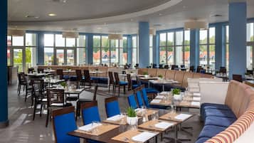 Een ruim restaurant met hoge plafonds, grote ramen en diverse zitmogelijkheden, zoals tafels en zitbanken.