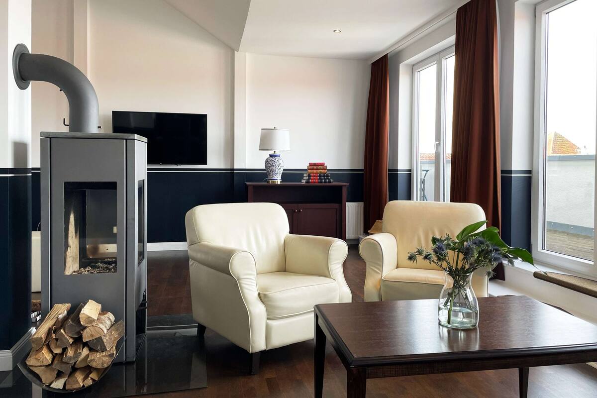 Un salon moderne avec une cheminée, deux fauteuils en cuir, une table basse en bois et une vue sur l’extérieur grâce à de grandes fenêtres.