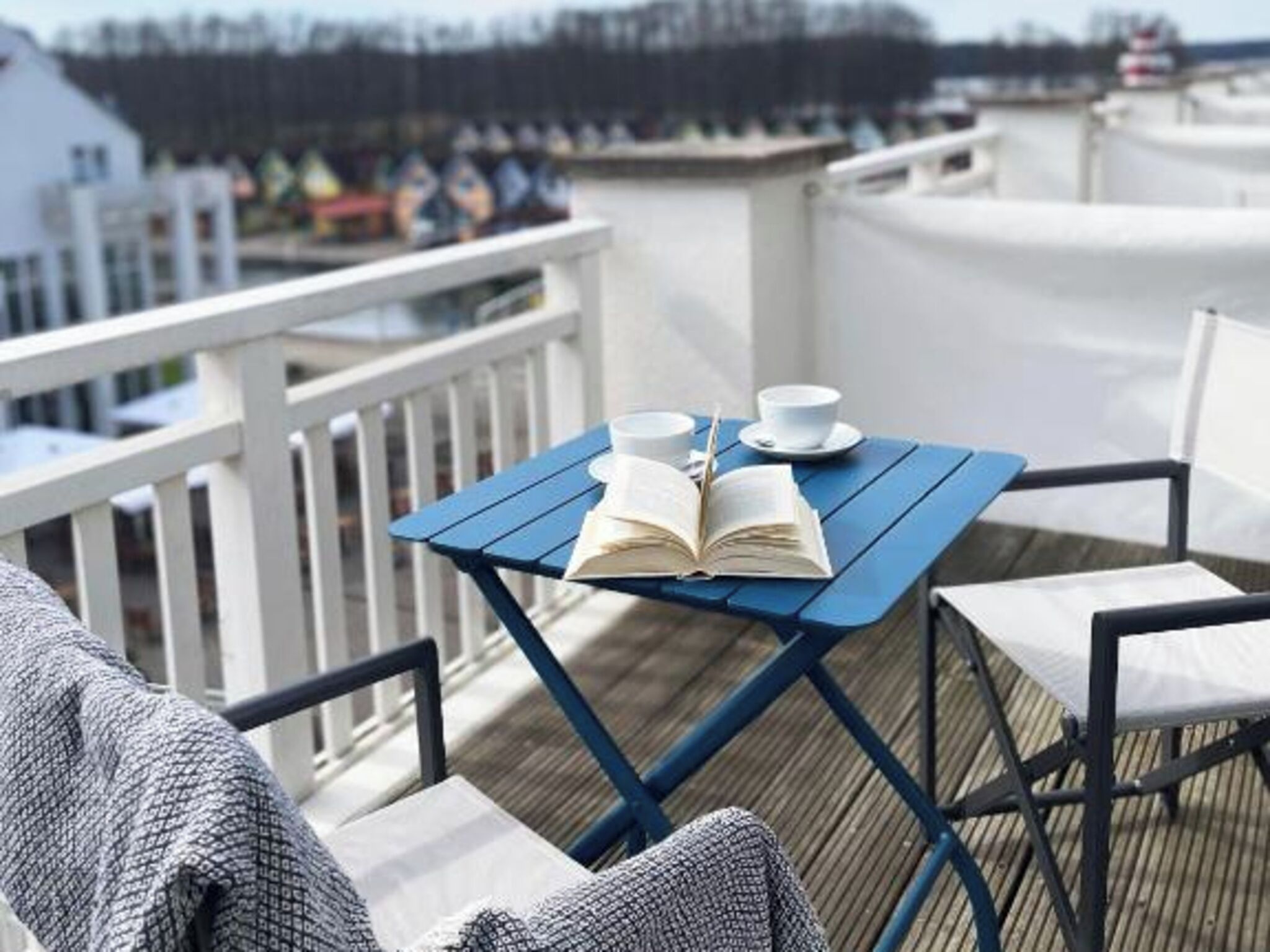 Una mesa azul con un libro abierto, dos tazas blancas y una silla sobre una cubierta de madera.