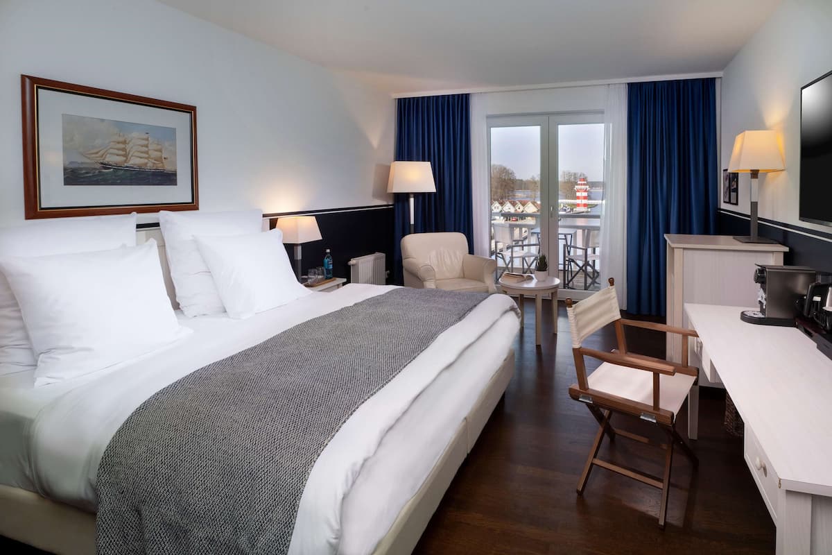 Une chambre d’hôtel avec un grand lit, un bureau, une chaise et une vue sur un balcon d’où l’on aperçoit un navire au loin.
