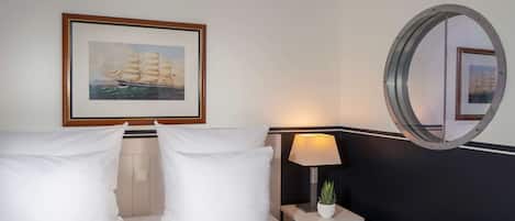 A hotel room with a bed, pillows, a nightstand with a lamp, a framed picture of a ship, and a round mirror.