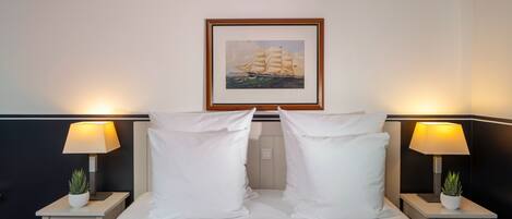Una cama bien hecha con almohadas blancas y un cuadro enmarcado de un barco velero sobre ella. Hay dos mesitas de noche con lámparas y pequeñas macetas.