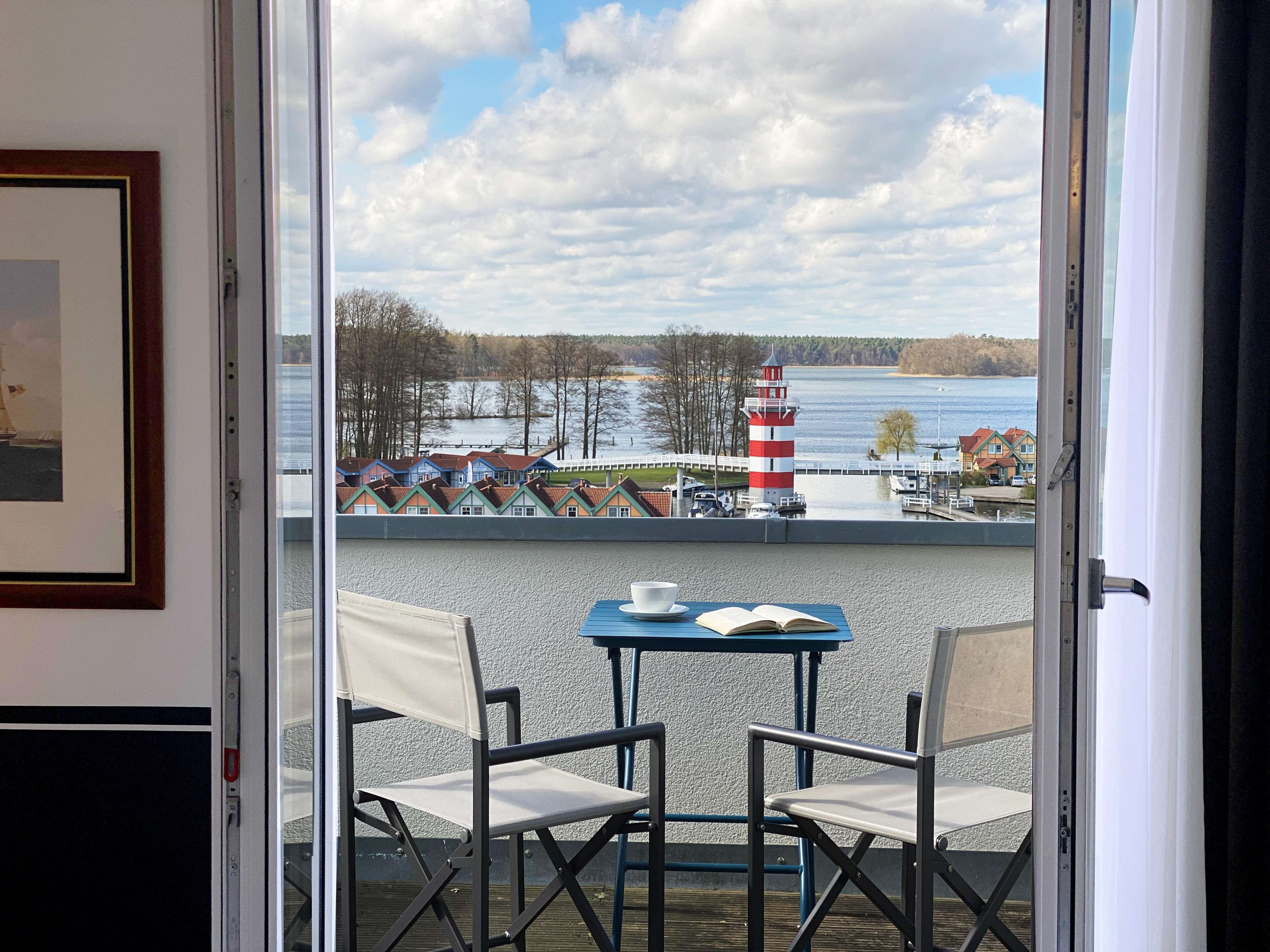 Een balkon met een tafel en twee stoelen, met uitzicht op een meer met huizen en een vuurtoren.