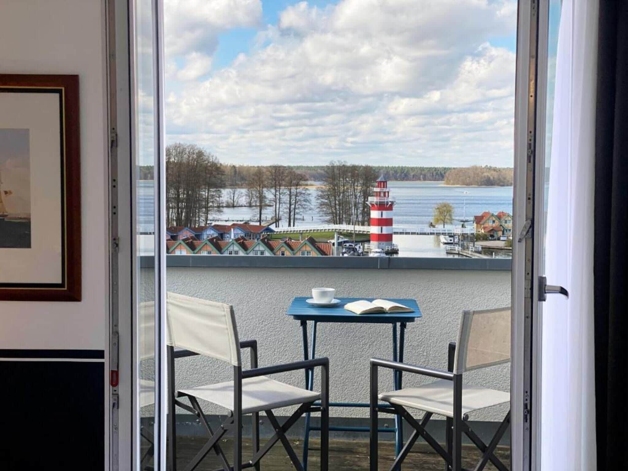 Ein Balkon mit zwei Stühlen, einem Tisch und Blick auf einen See mit einem Leuchtturm.
