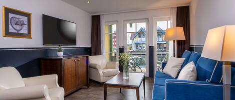 A modern hotel room with a flat-screen TV, a sofa, a chair, a wooden cabinet, and a view of colorful buildings outside.