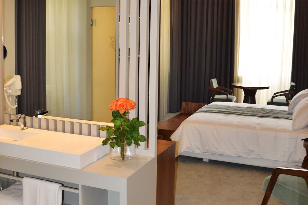 A modern hotel room with a large bed, a separate bathroom with a vanity, and a seating area with two chairs and a table.