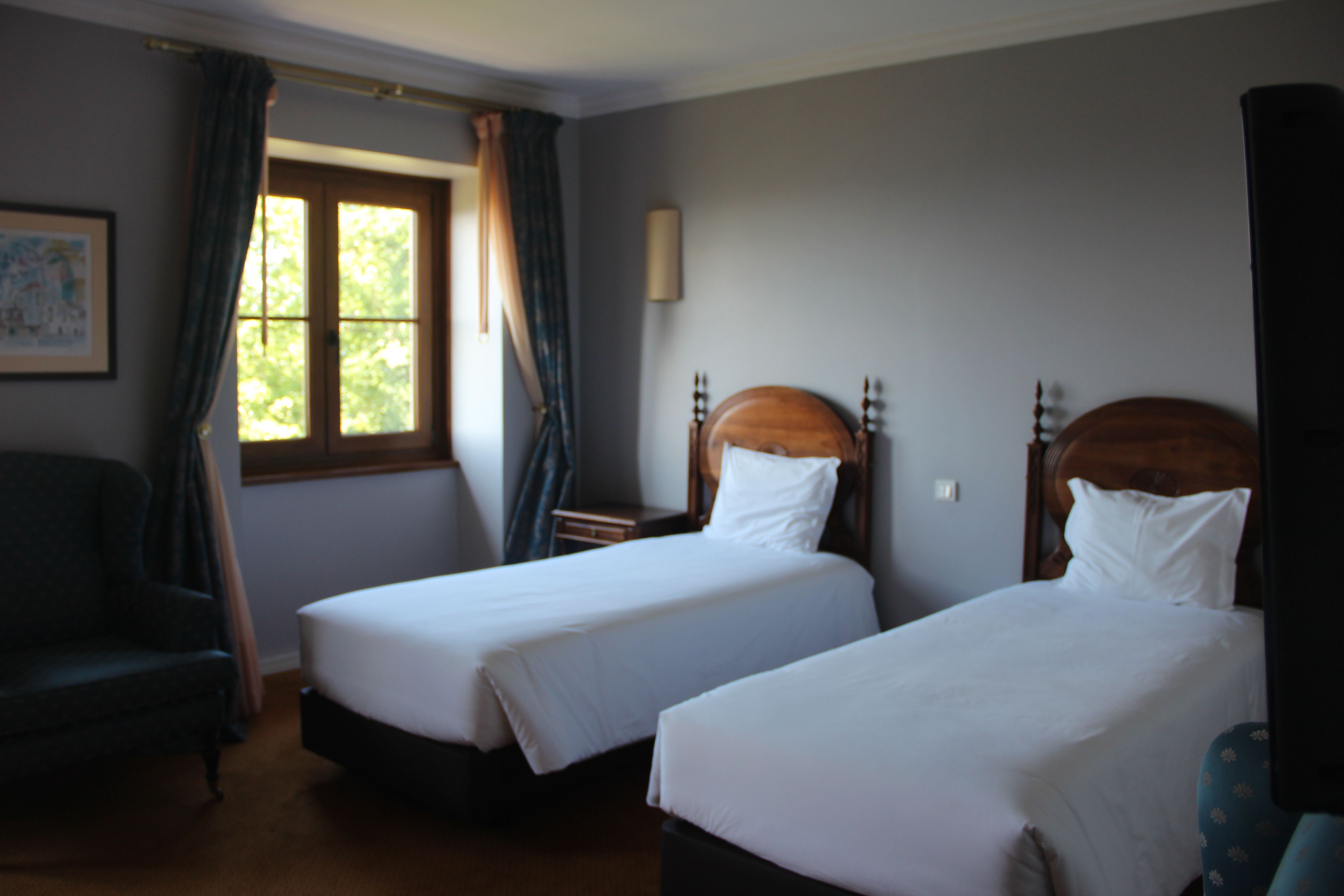 A hotel room with two single beds, a chair, a window with curtains, and a painting on the wall.