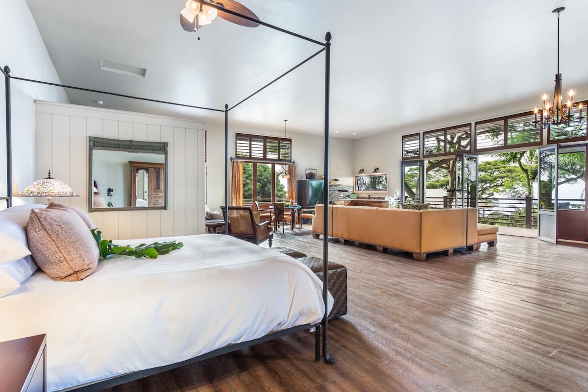 A spacious bedroom with a large bed, a sitting area with a sofa and chairs, and a view of the outdoors through large windows.