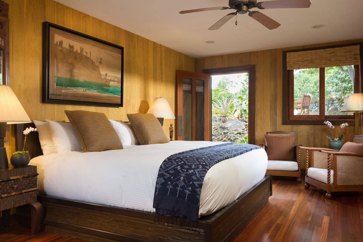 A bedroom with a large bed, wooden walls, a ceiling fan, a window with a view of greenery, and a framed picture on the wall.