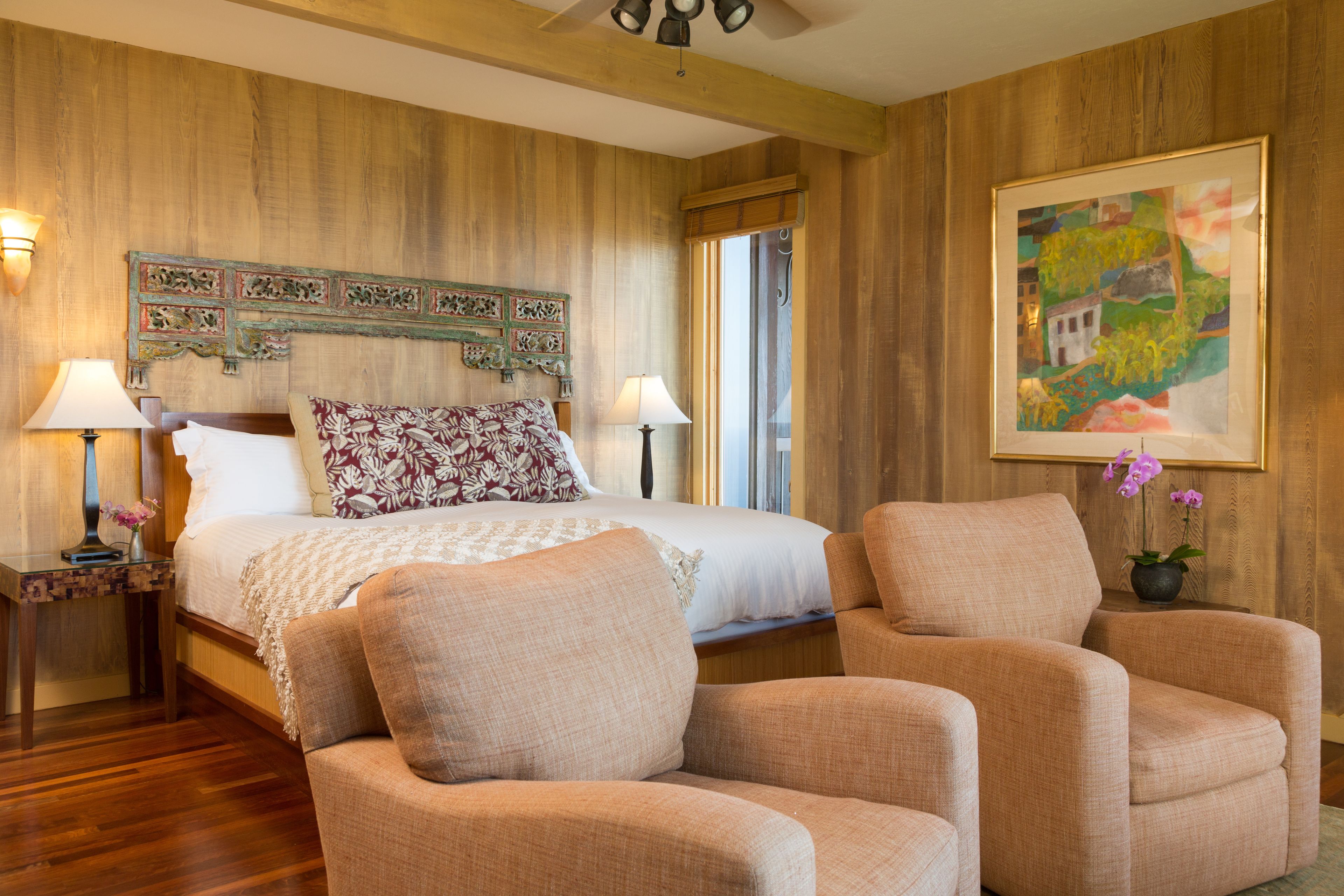A hotel room with a wooden headboard, a large bed with patterned pillows, two bedside lamps, and a framed painting on the wall.