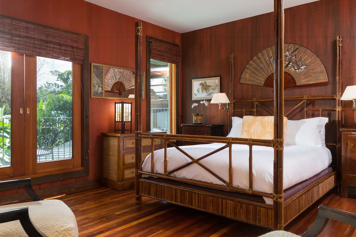 A four-poster bed with a wooden frame, a nightstand with a lamp, and a large window with a view of greenery.