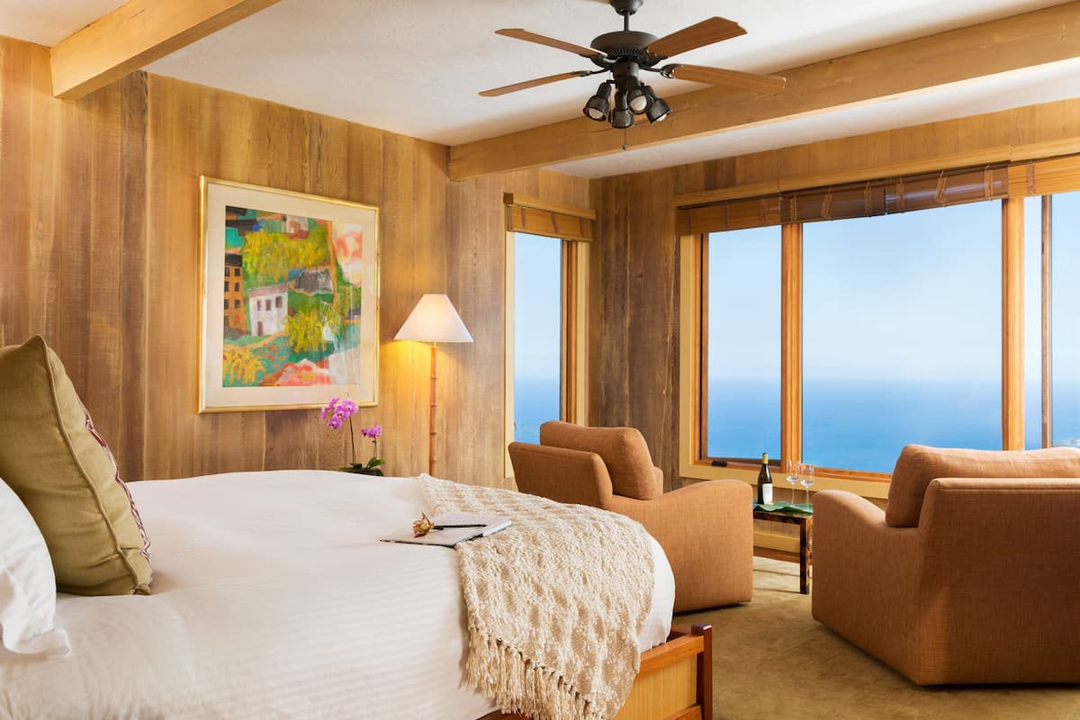 A hotel room with a large bed, a ceiling fan, a painting, a view of the ocean, and a seating area with a table.