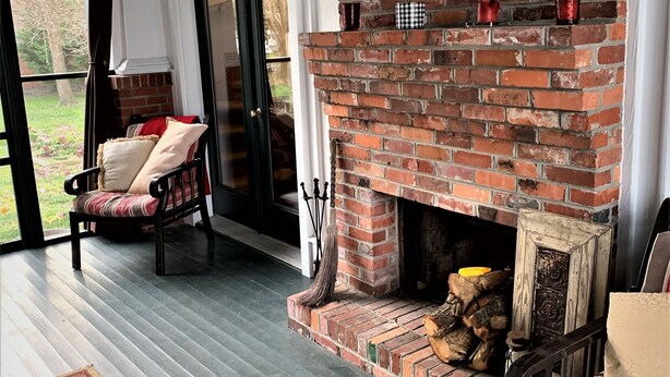 A cozy room with a brick fireplace, a comfortable chair, a rug, and a view through a glass door. - Cameron House Inn (Manteo)