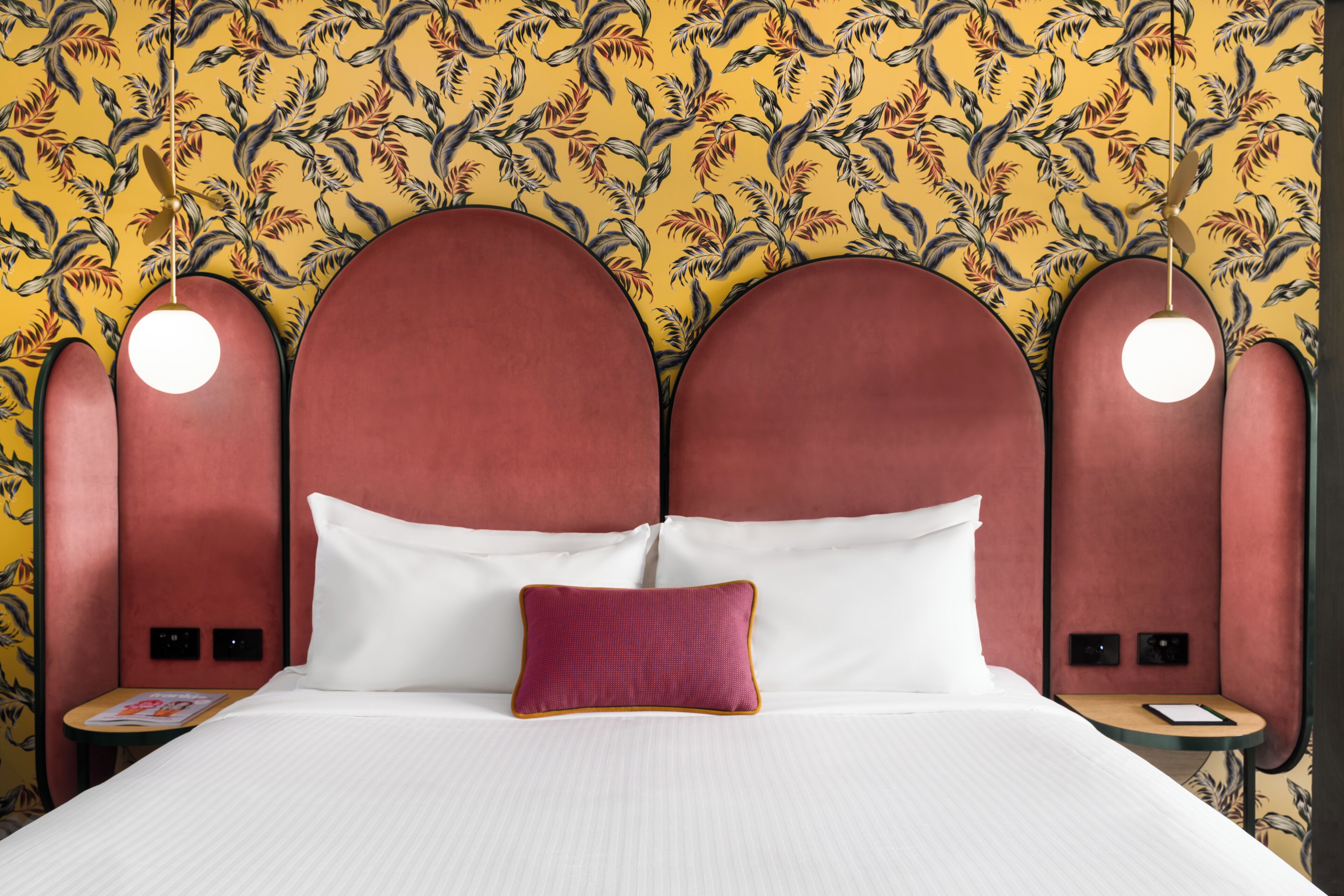 Una habitación de hotel con cama, mesita de noche, lámpara de lectura y un muro decorado con un diseño de hojas.