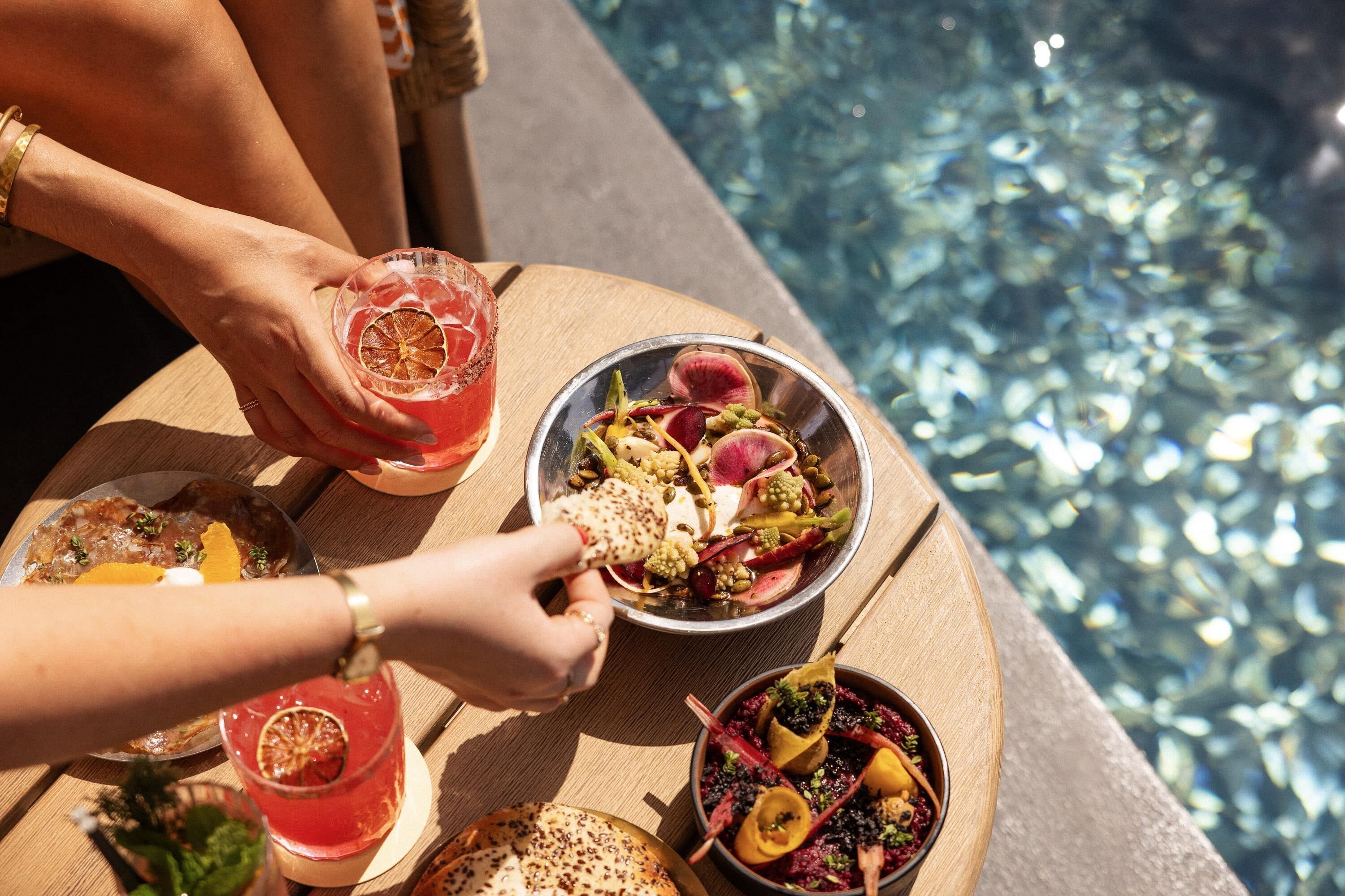 A dining table with various dishes and drinks, a pool in the background, and a person holding a drink.