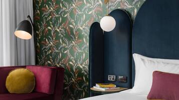 A hotel room with a dark blue headboard, a red sofa with a yellow pillow, a round table with books, and a wall with a leafy pattern.