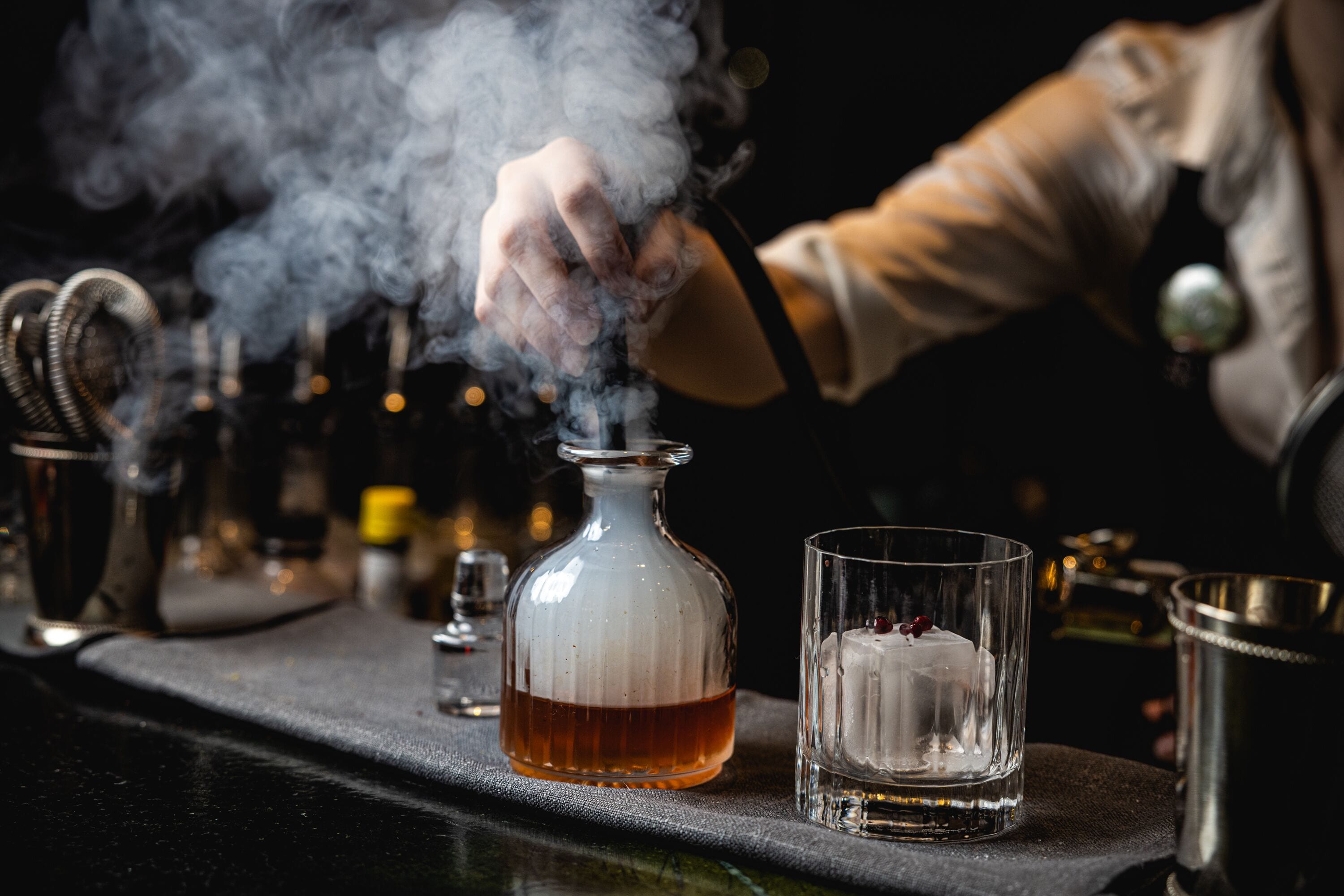 A bar setting with a glass decanter, a glass with a candle, and a metal shaker on a bar counter.